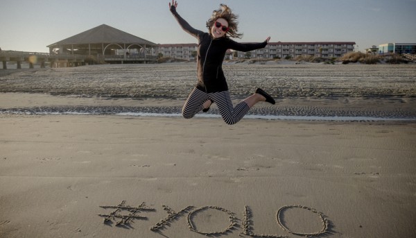 A woman jumping over the English slang term "yolo" that she has written on the beach.