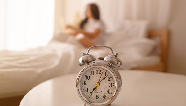 an alarm clock on a table with a woman in bed in the background, consistency is important for a good routine